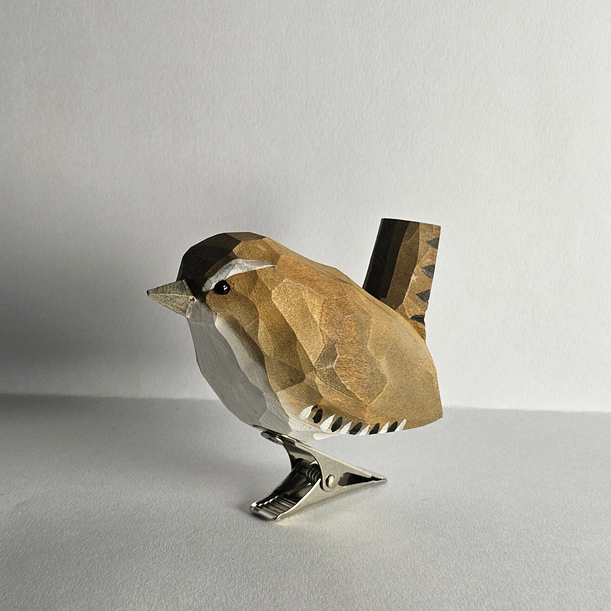 Side view of a hand-carved, hand-painted wooden Winter Wren bird clip-on ornament mounted on a metal spring clip, resting on a plain white surface.