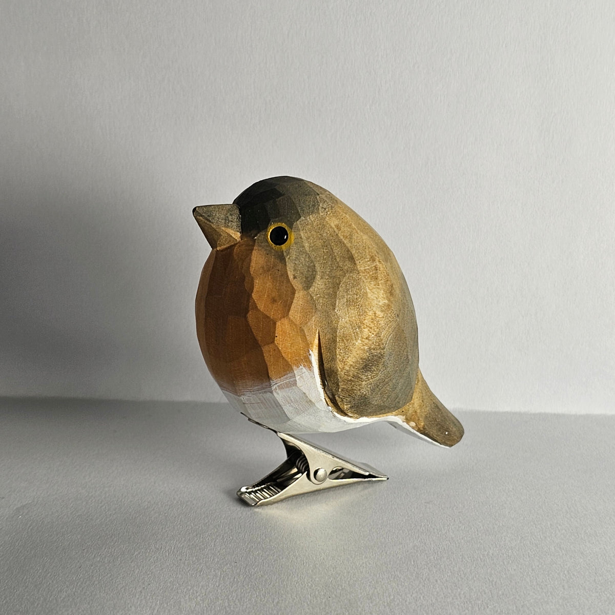 A close-up side-profile shot of a hand-carved wooden European Robin clip-on ornament resting on a white surface, featuring a warm olive-brown back and wing plumage, a soft orange-red face and breast, a light grey belly, and a silver metal clip attached underneath.