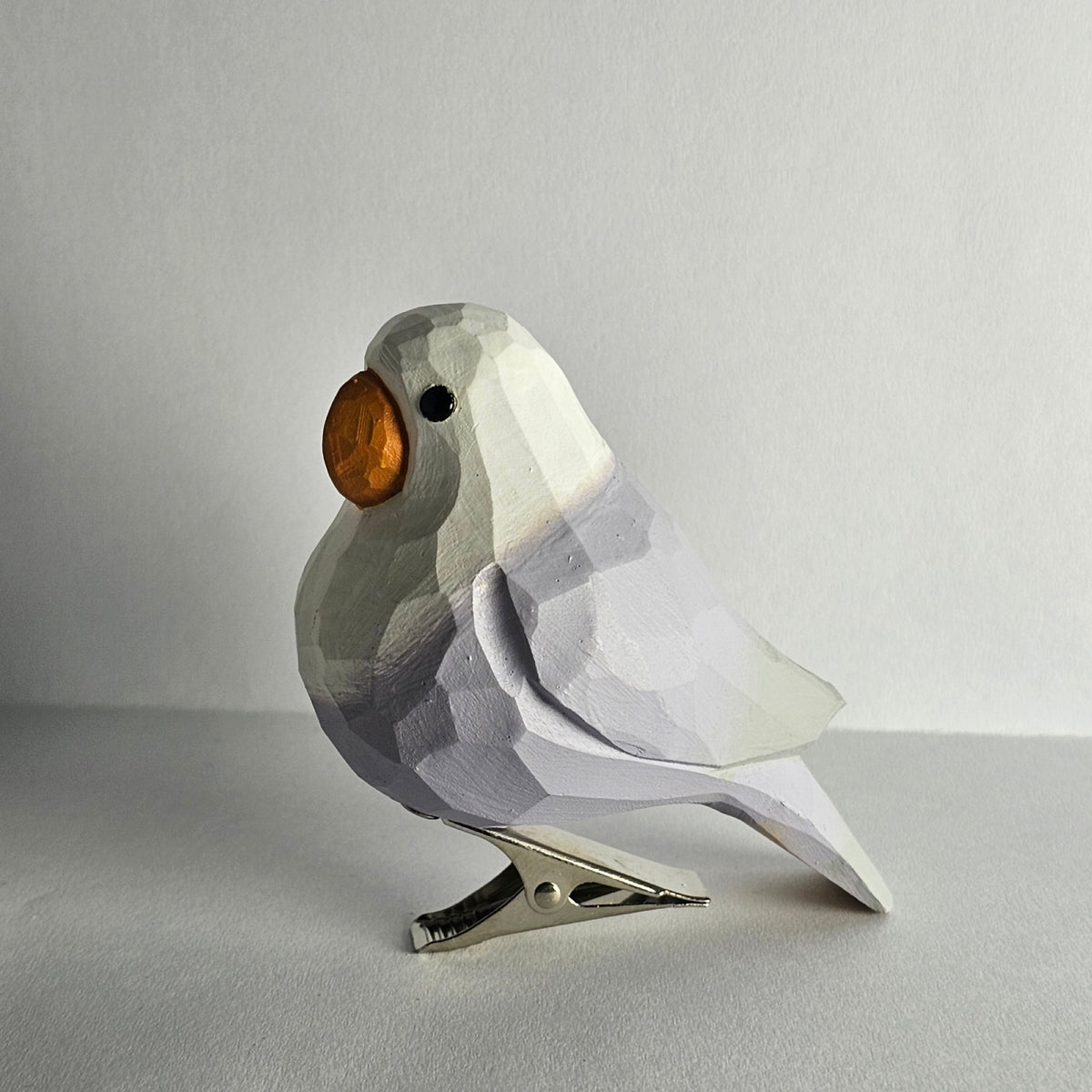 A close-up side-profile photo of a hand-carved wooden purple Lovebird clip-on bird ornament resting on a plain light grey surface, featuring pale lavender and white painted plumage, a bright orange beak, a dark eye, and a silver metal clip attached underneath.