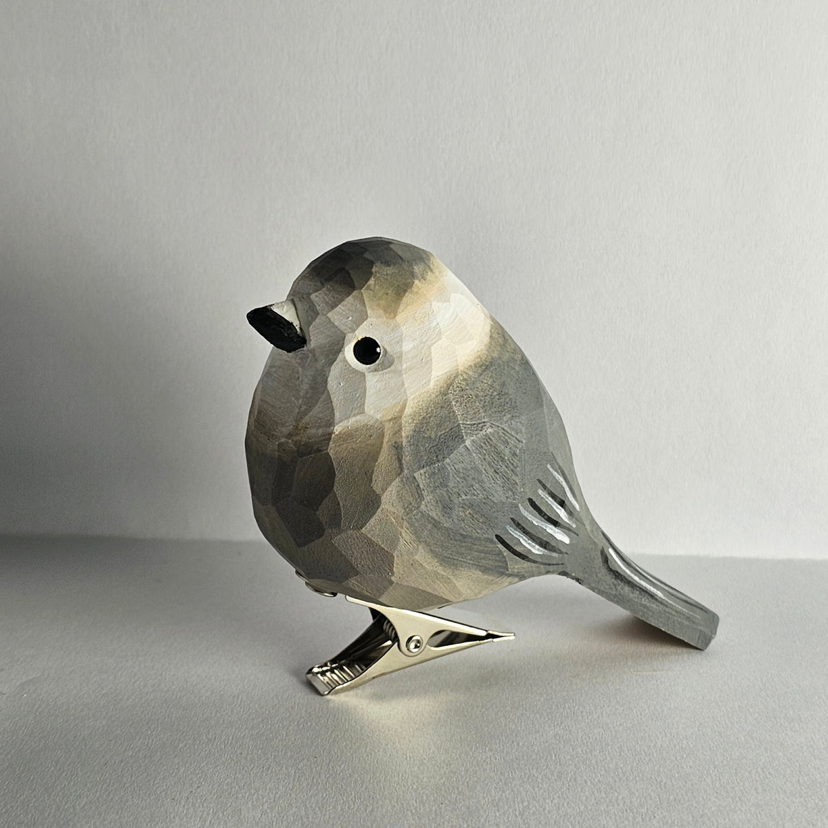A side-profile shot of a hand-carved wooden Canada Jay clip-on ornament resting on a plain light grey surface, featuring textured grey plumage, a white forehead and throat, a dark grey cap, and a silver metal clip attached underneath.