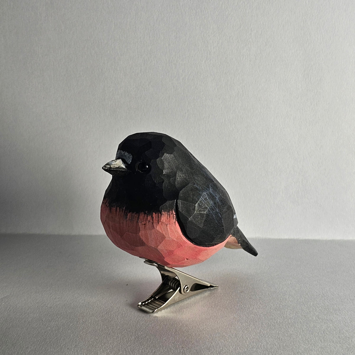 A close-up side-profile shot of a hand-carved wooden Pink Robin clip-on ornament resting on a plain light grey surface, featuring a jet-black head, back, and wings, a striking rose-pink breast, a pale grey belly, and a silver metal clip attached underneath.
