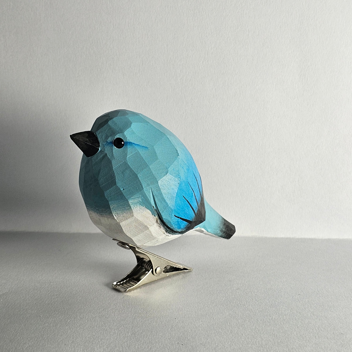 A hand-carved blue wooden bird ornament shown in profile against a light background, featuring a painted white breast, a dark beak, and a silver clip attached to its underside.