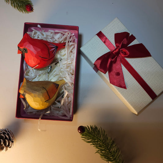 Hand-carved wooden male and female cardinal clip-on ornaments resting inside an open red gift box with paper shred, next to a matching gift box lid tied with a red ribbon and festive pine accents.