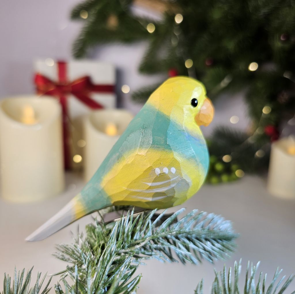 A hand-carved wooden lovebird clip-on ornament shown in profile, featuring a bright yellow cap, a soft turquoise-blue and light-green body with striped wing accents, an orange beak, and clipped onto an evergreen pine branch with lit candles in the background.