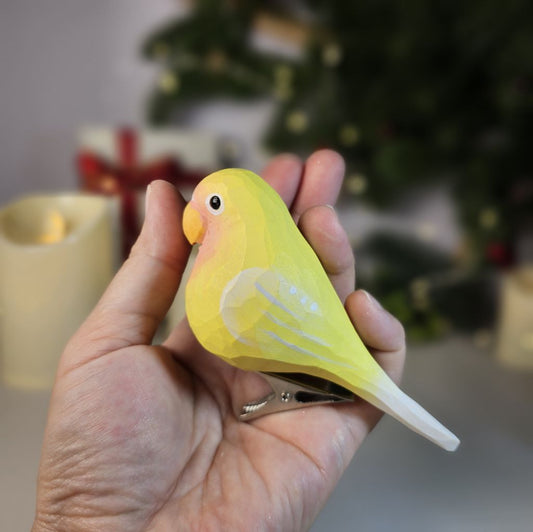 A hand-carved yellow wooden lovebird clip-on ornament held resting in an open palm, featuring a light pink face, pastel yellow body, soft white wing accents, and a silver clip attached underneath.