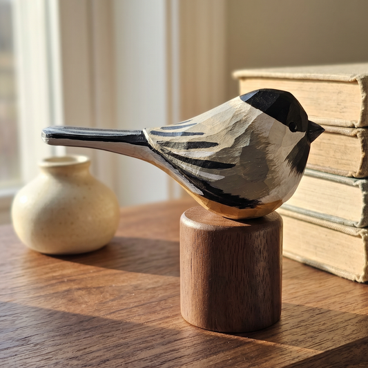 A hand-carved wooden Black-capped Chickadee figurine with black, white, and gray painted details, perched on a cylindrical wooden block on a wooden table. In the background, there is a small beige ceramic pot and a stack of vintage books near a window.