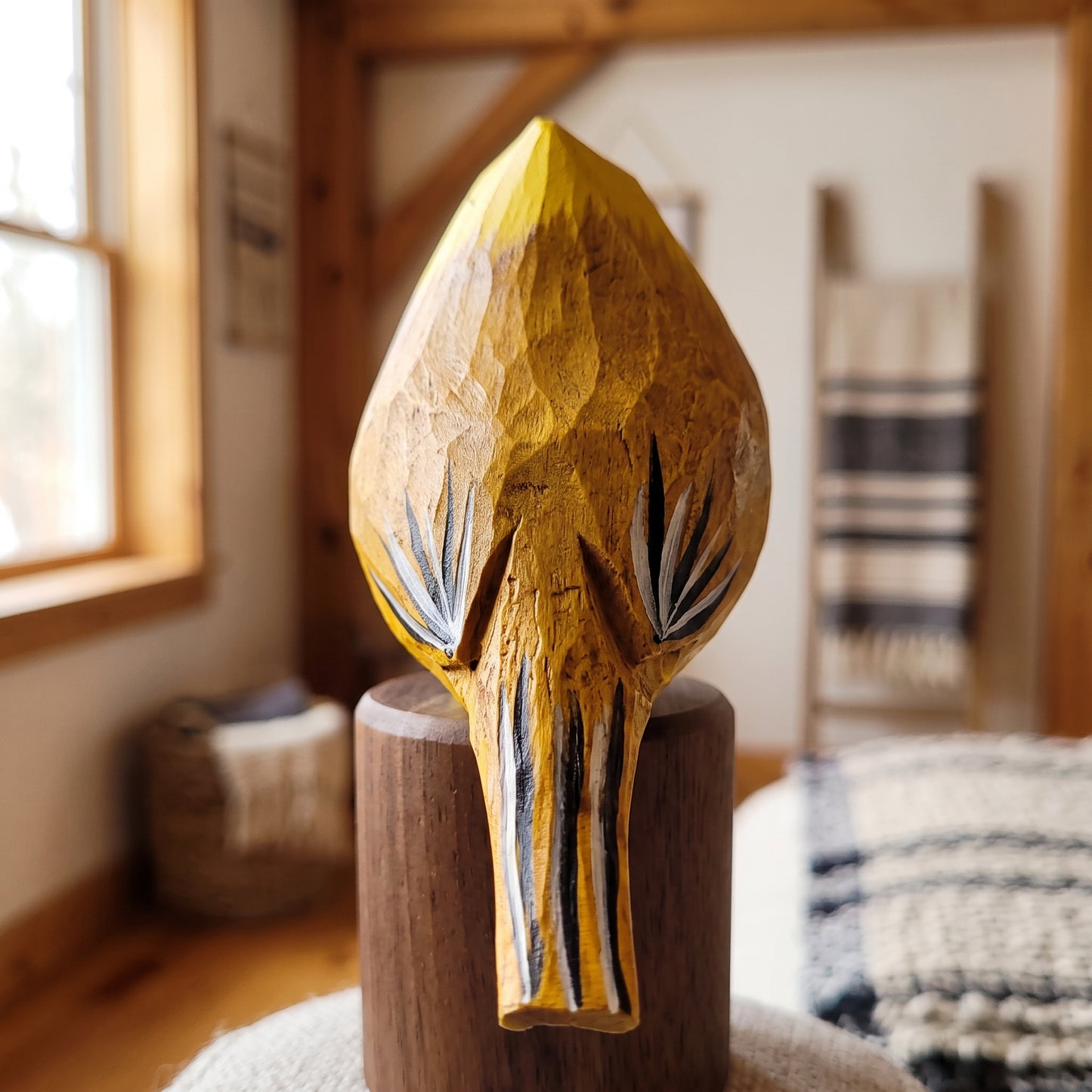 A hand-carved wooden Yellow Cardinal sculpture of a stylized animal head, likely a bison or antelope, in warm natural wood tones with black and white painted details, displayed on a wooden pedestal in a rustic indoor setting.