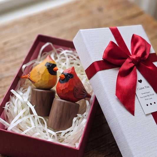A gift box containing two wooden cardinal figurines, one orange-yellow and one red, displayed next to its white lid wrapped with a red ribbon on a rustic wooden table.