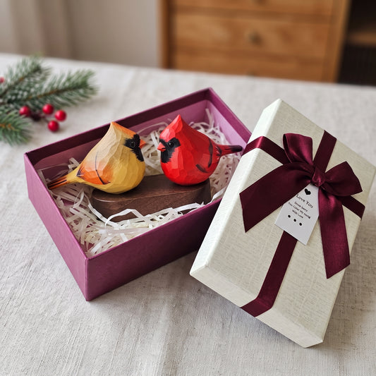 A gift box set containing two wooden cardinal figurines, one yellow and one red, displayed inside an open magenta box next to its cream-colored lid wrapped in a burgundy ribbon.