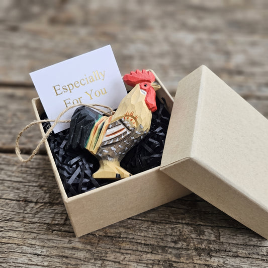 A handcrafted wooden rooster ornament nestled in black shredded paper inside a kraft gift box, next to a white card that reads 'Especially For You'. The box is placed on a rustic wooden surface.