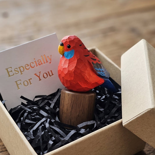 A small Crimson rosella figurine with blue and yellow accents sits on a wooden stand inside a kraft gift box with black shredded paper and a card that reads 'Especially For You,' placed on a wooden table.