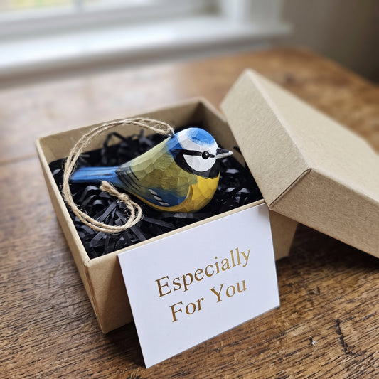 A small Eurasian blue tit-shaped ornament in blue, yellow, and black sits in a kraft gift box filled with black shredded paper, with a white card reading 'Especially For You' in front. The box is open on a wooden table near a window.