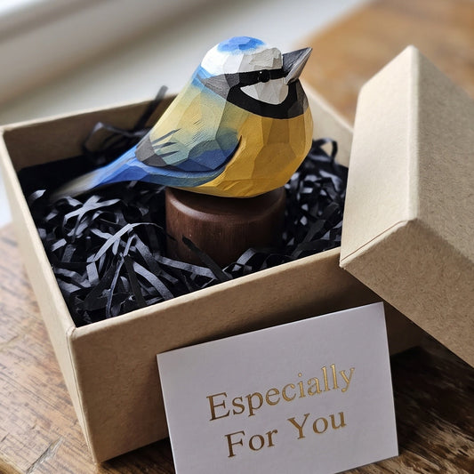 A small handcrafted Eurasian blue tit figurine with yellow, blue, black, and white geometric panels sits in a brown kraft gift box filled with black paper shred. A white card reading 'Especially For You' is placed in front, and the box rests on a rustic wooden table near a window.