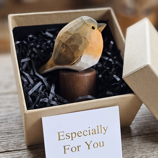 A small carved European robin figurine in a kraft gift box with black crinkle paper, with the lid leaning against the box and a card reading 'Especially For You' in front. The background is a softly blurred rustic wooden setting.