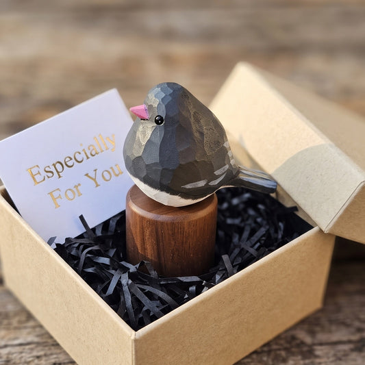 A small Junco figurine with a dark gray faceted body, white underbelly, and pink beak perched on a wooden stand inside a kraft gift box with black shredded paper and a card that reads 'Especially For You'.