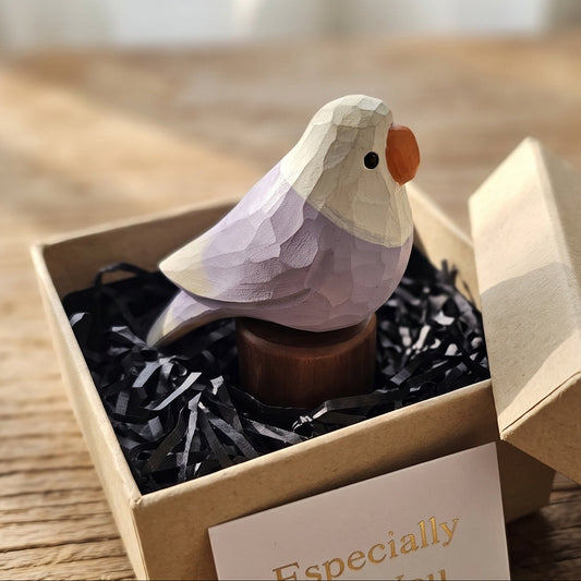 A geometric Lovebirds wooden ornament with a lavender body, cream head, and orange beak, displayed in a kraft gift box with black shredded paper on a wooden surface.