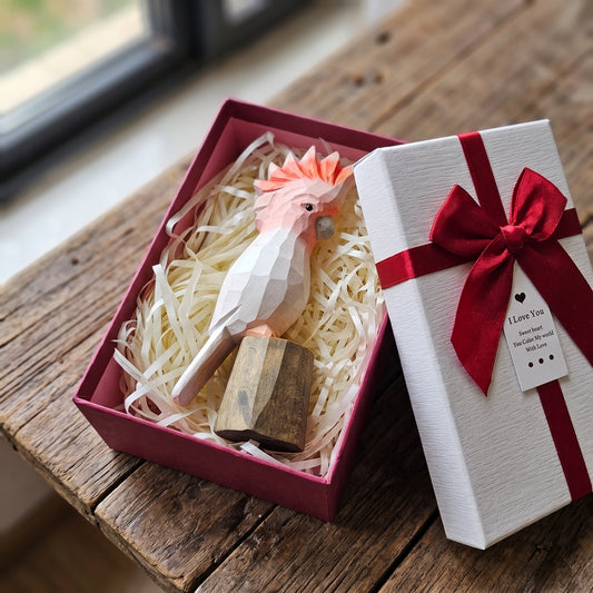 Hand-carved wooden Pink Cockatoo bird figurine resting inside an open gift box with shredded paper filler, next to a white gift box lid tied with a red ribbon.