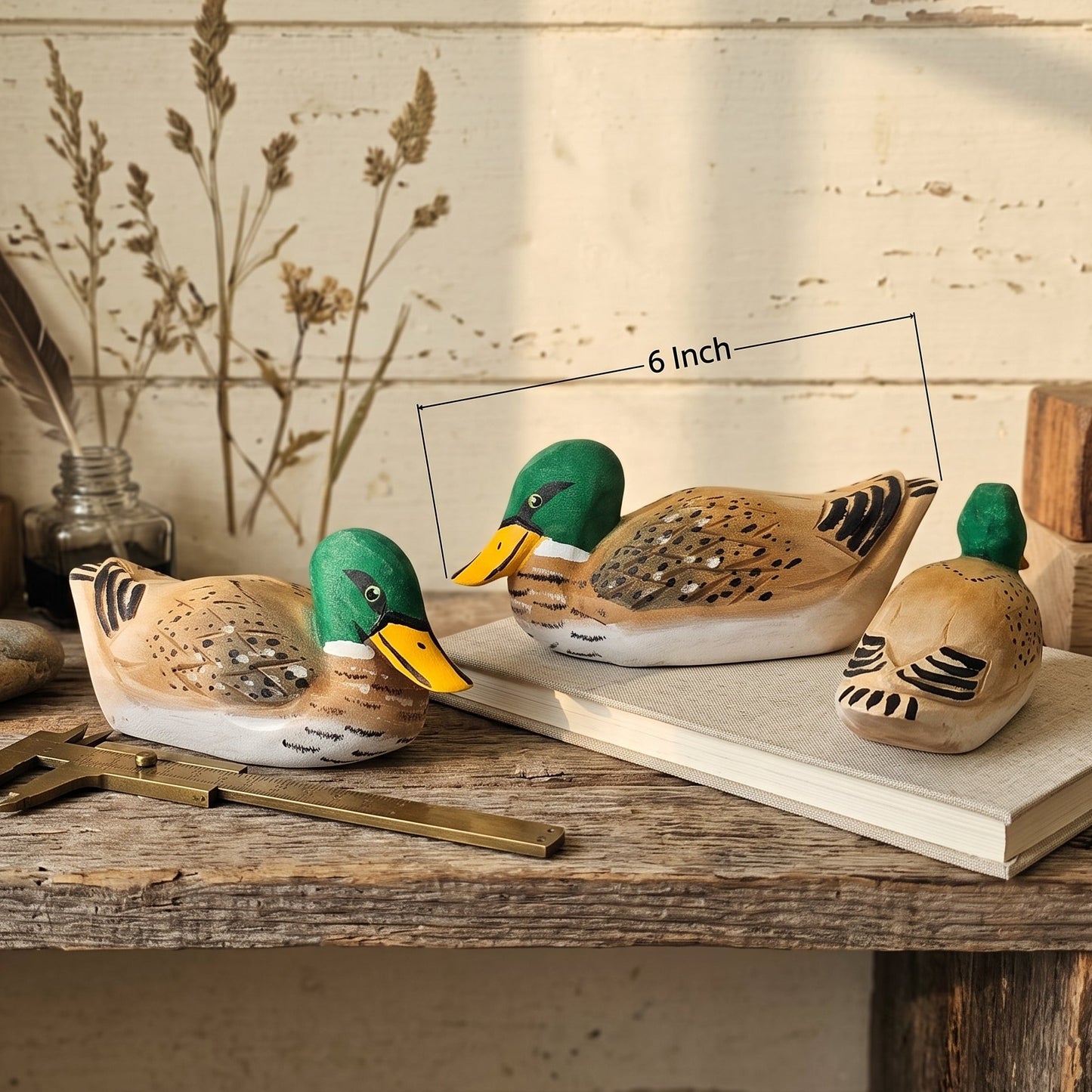 Three hand-painted wooden mallard ducks displayed on a rustic wooden table with a vintage brass caliper and a size indicator line showing 6 inch length. The background features a distressed cream-colored wooden wall with a feather in a glass bottle.