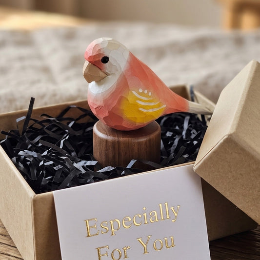 A small painted Lovebird figurine perched on a wooden base, displayed inside an open kraft gift box with black shredded paper and a card that reads 'Especially For You.' The box sits on a wooden surface in a softly lit indoor setting.