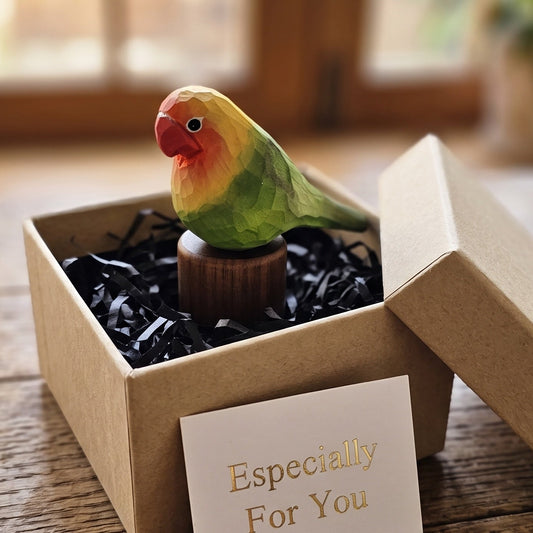 A small wooden lovebird figurine perched on a wooden base, presented inside an open kraft gift box with black shredded paper and a card that reads 'Especially For You' on a rustic wooden table.
