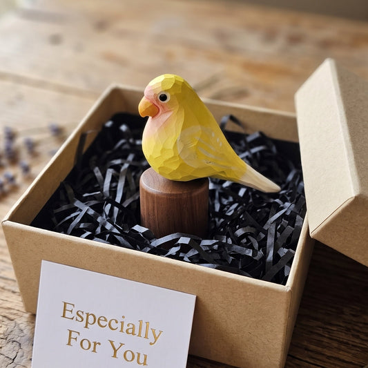 A small yellow Lovebird figurine with pink and orange details sits on a wooden stand inside a kraft gift box filled with black paper shred. A white card reading 'Especially For You' is placed in front, and the box lid rests beside it on a rustic wooden table.