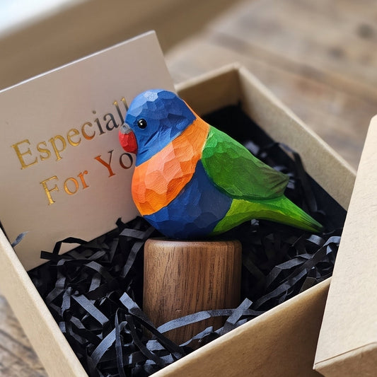 A small colorful Rainbow lorikeet figurine with a faceted geometric design sits in a brown kraft gift box filled with black paper shred, next to a card that reads 'Especially For You'. The box is placed on a rustic wooden table near a window.
