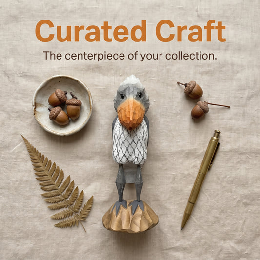 A styled flat-lay featuring a carved Shoebill figurine at the center, surrounded by acorns in a small dish, a dried fern leaf, a gold pen, and text reading 'Curated Craft' on a beige fabric background.