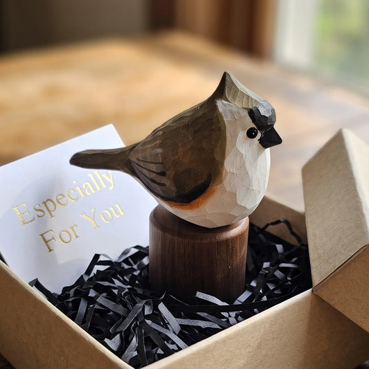 A handcrafted wooden Tufted Titmouse figurine with a white, gray, and brown body sits on a wooden base inside a kraft gift box filled with black shredded paper and a card that reads 'Especially For You,' placed on a rustic wooden table.