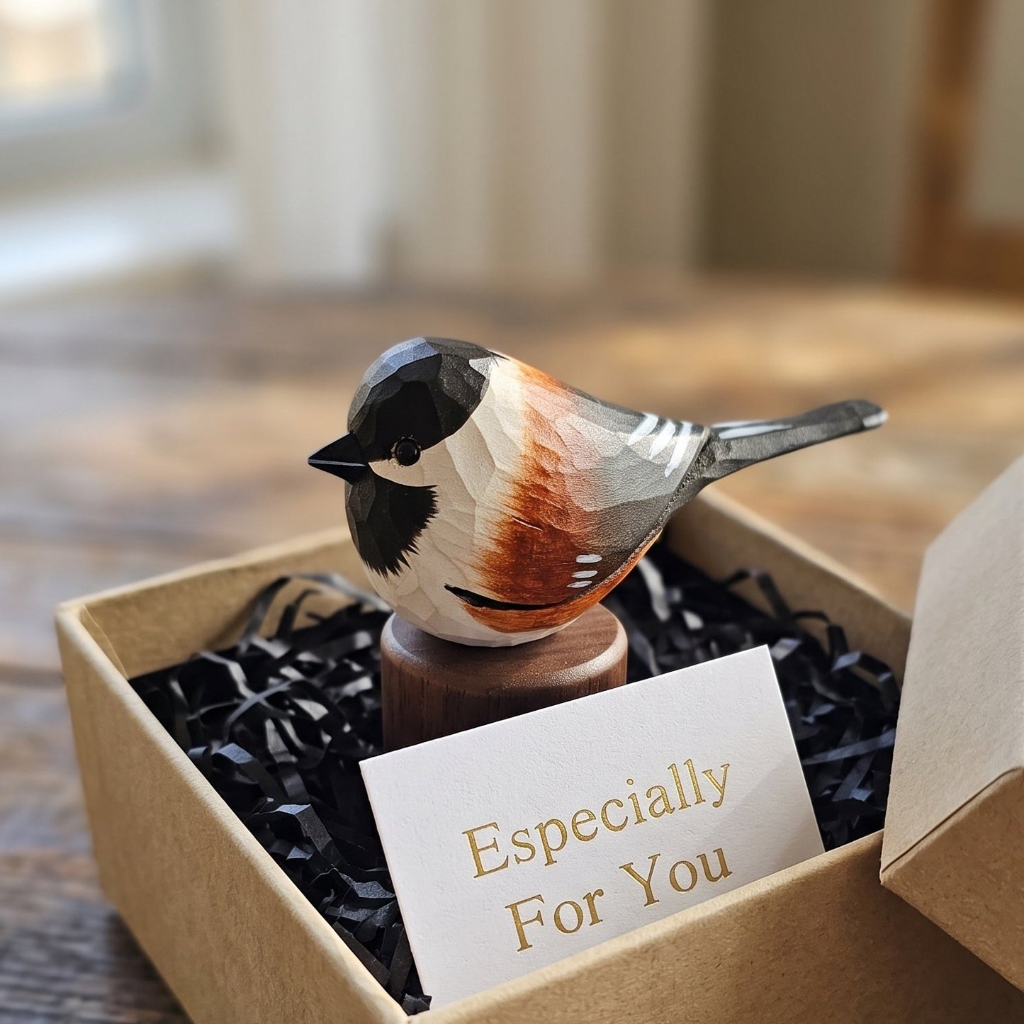 A small Chestnut-backed chickadee figurine perched on a wooden stand, presented in an open kraft gift box with black crinkle paper filler and a card that reads 'Especially For You.' The bird has a black head, white body, gray wings, and rust-orange back.
