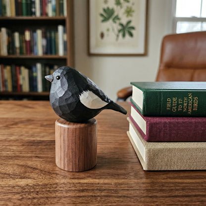 A carved wooden Lark bunting figurine with a faceted geometric pattern, perched on a small cylindrical wooden base. The bird is black with white accents, set on a wooden table next to a stack of books in a cozy, softly lit room with a bookshelf in the background.