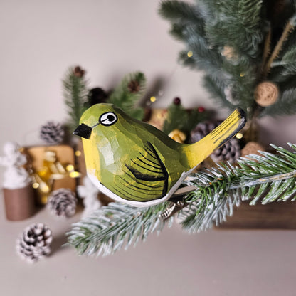 A close-up side-profile photo of a hand-carved wooden White-Eye clip-on bird ornament clipped onto a faux evergreen branch, featuring vibrant yellow-green plumage, a white belly, a black bill, and a signature white eye-ring, set against a blurred holiday backdrop with pinecones and fairy lights.