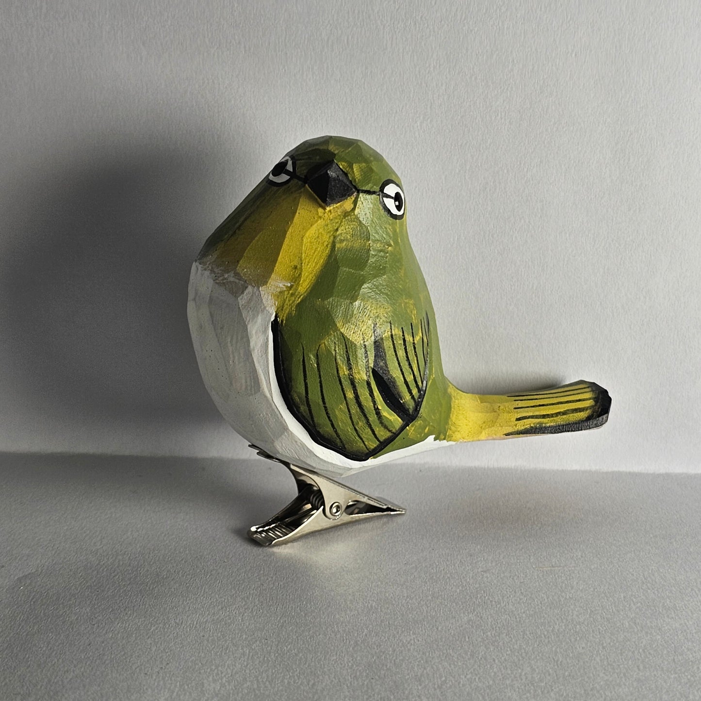 A close-up side-profile photo of a hand-carved wooden White-Eye bird clip-on ornament resting on a plain light grey surface, featuring vibrant yellow-green head and wing plumage, a white breast and belly, a black beak, a distinct white ring around its eye, and a silver metal clip attached underneath.