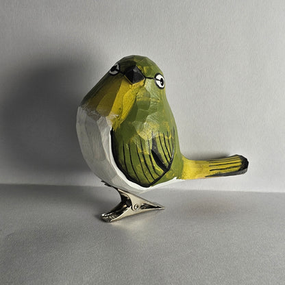 A close-up side-profile photo of a hand-carved wooden White-Eye bird clip-on ornament resting on a plain light grey surface, featuring vibrant yellow-green head and wing plumage, a white breast and belly, a black beak, a distinct white ring around its eye, and a silver metal clip attached underneath.