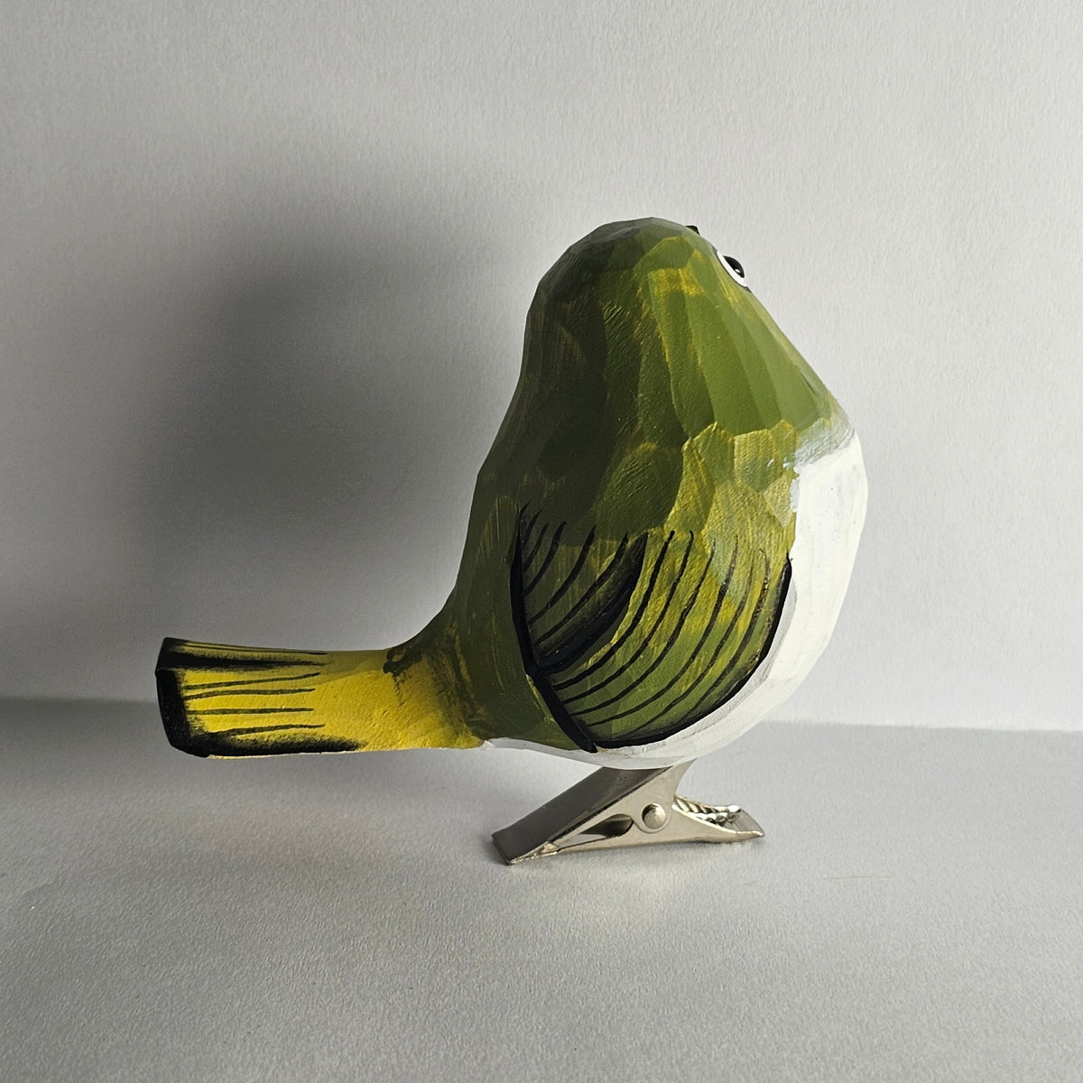 A close-up side-profile photo of a hand-carved wooden White-Eye clip-on bird ornament shown from behind, featuring green and yellow-painted upper plumage with carved feather grooves, a white underbelly, a yellow tail tip, a subtle eye-ring detail, and a silver metal clip attached underneath against a light grey background.