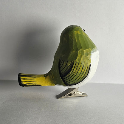 A close-up side-profile photo of a hand-carved wooden White-Eye clip-on bird ornament shown from behind, featuring green and yellow-painted upper plumage with carved feather grooves, a white underbelly, a yellow tail tip, a subtle eye-ring detail, and a silver metal clip attached underneath against a light grey background.