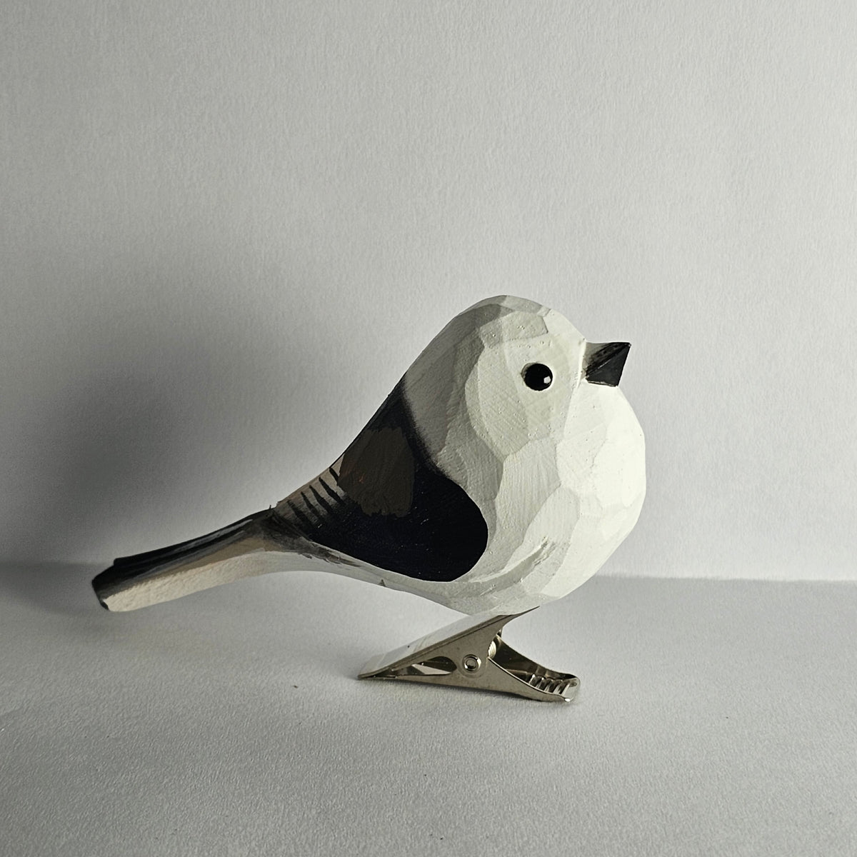 A close-up side-profile photo of a hand-carved wooden Long-Tailed Tit clip-on bird ornament resting on a plain light grey surface, featuring a white head and body with black wing markings, a long black tail, a small black eye, a tiny beak, and a silver metal clip attached underneath.