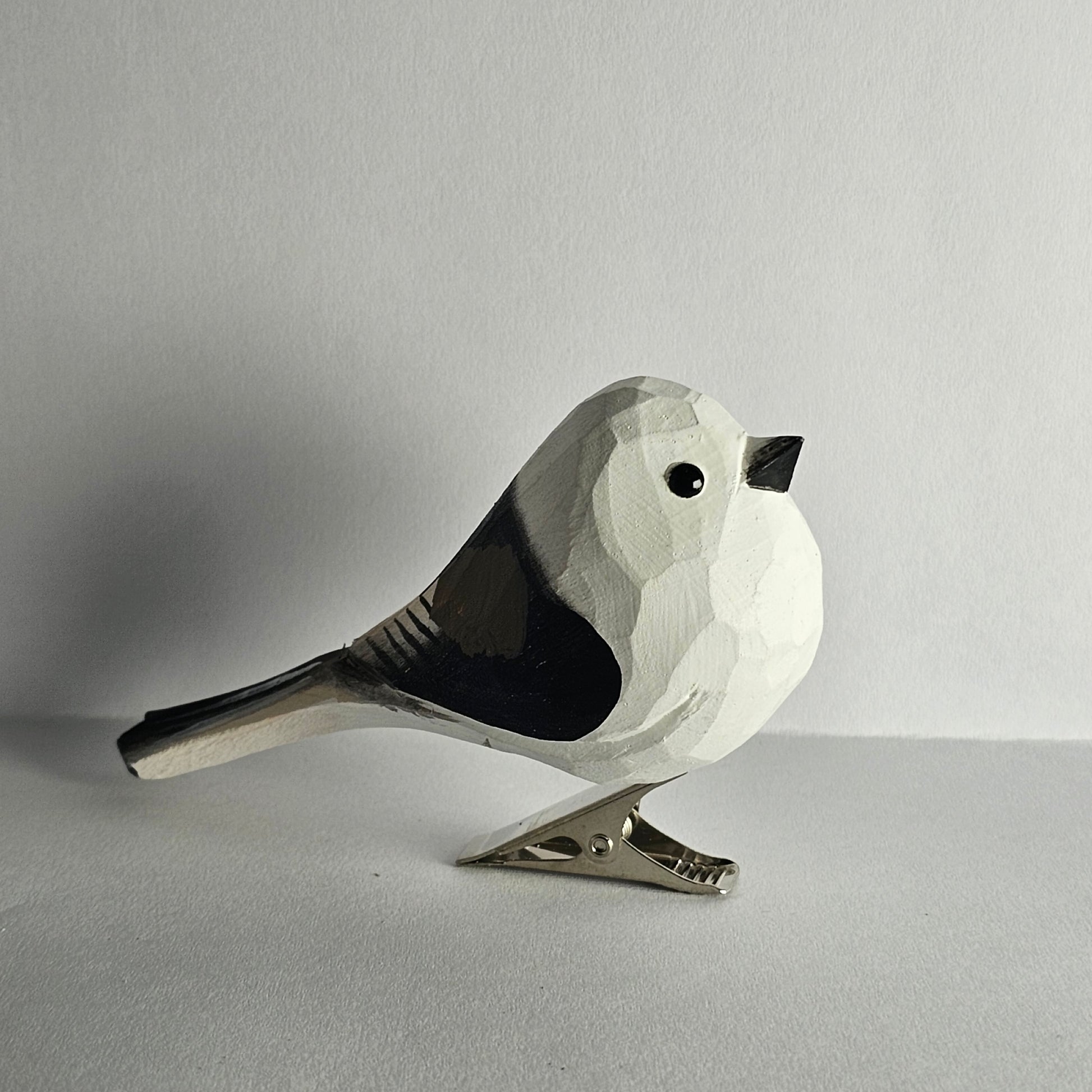 A close-up side-profile photo of a hand-carved wooden Long-Tailed Tit clip-on bird ornament resting on a plain light grey surface, featuring a white head and body with black wing markings, a long black tail, a small black eye, a tiny beak, and a silver metal clip attached underneath.