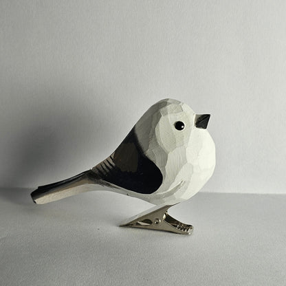 A close-up side-profile photo of a hand-carved wooden Long-Tailed Tit clip-on bird ornament resting on a plain light grey surface, featuring a white head and body with black wing markings, a long black tail, a small black eye, a tiny beak, and a silver metal clip attached underneath.