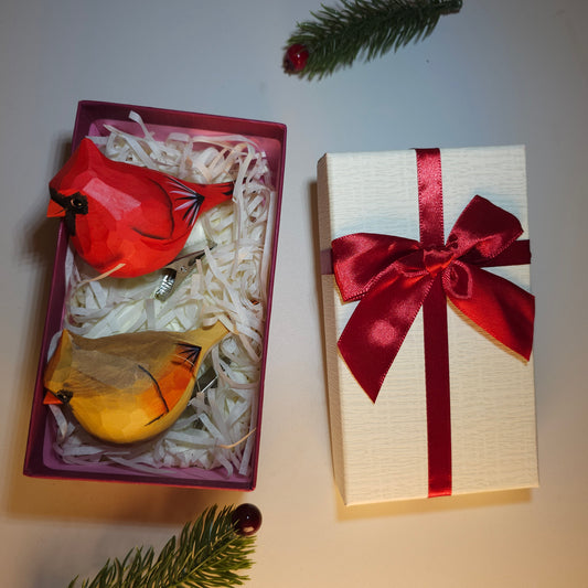 Hand-carved wooden male and female cardinal clip-on ornaments resting in a gift box filled with shred, alongside a white textured lid tied with a red satin bow.