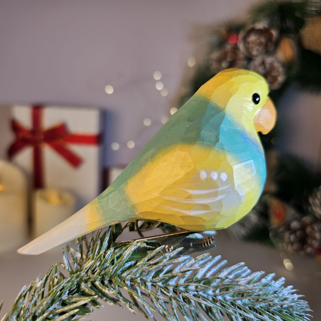 A hand-carved wooden lovebird clip-on ornament shown in profile, featuring a pastel yellow cap, a light green body with turquoise and white wing accents, a dark eye, a light orange beak, and clipped onto an evergreen pine branch with soft holiday bokeh lights in the background.