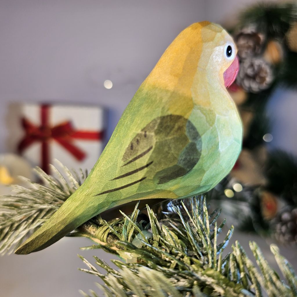 A hand-carved yellow wooden lovebird clip-on ornament shown in profile, featuring a pastel green body with soft yellow and pink accents, a black eye, a light pink beak, and clipped onto an evergreen pine branch with soft holiday bokeh lights in the background.