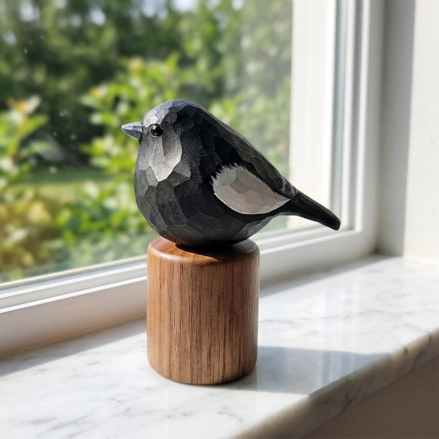A carved wooden Lark bunting figurine with a geometric, faceted design, perched on a cylindrical wooden base. The bird is positioned on a white marble windowsill with a soft-focus green garden background visible through the window.