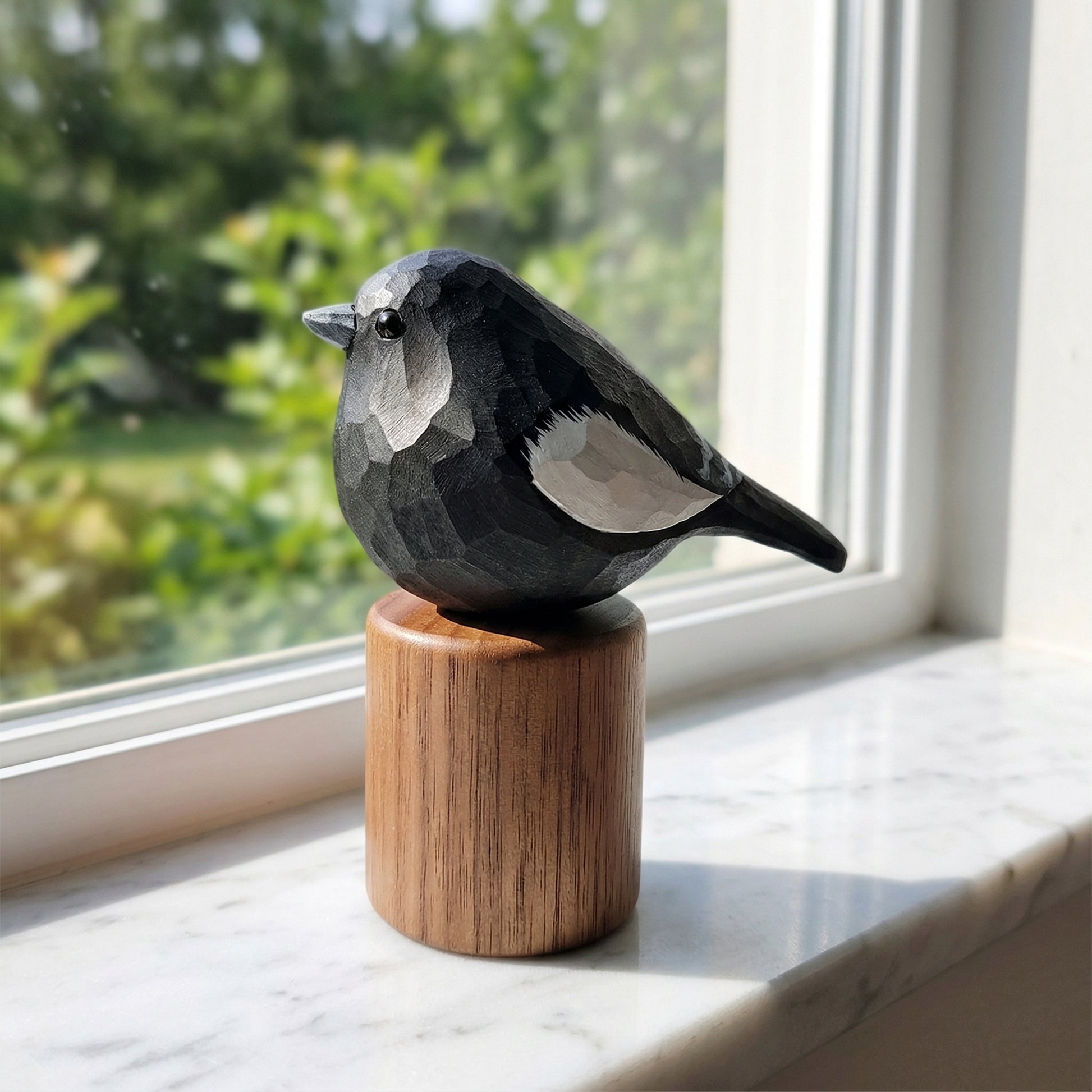 A carved wooden Lark bunting figurine with a geometric, faceted design, perched on a cylindrical wooden base. The bird is positioned on a white marble windowsill with a soft-focus green garden background visible through the window.