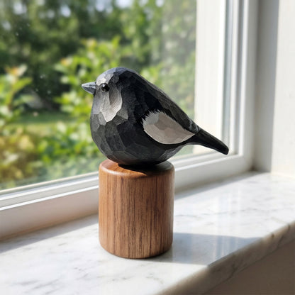 A carved wooden Lark bunting figurine with a geometric, faceted design, perched on a cylindrical wooden base. The bird is positioned on a white marble windowsill with a soft-focus green garden background visible through the window.