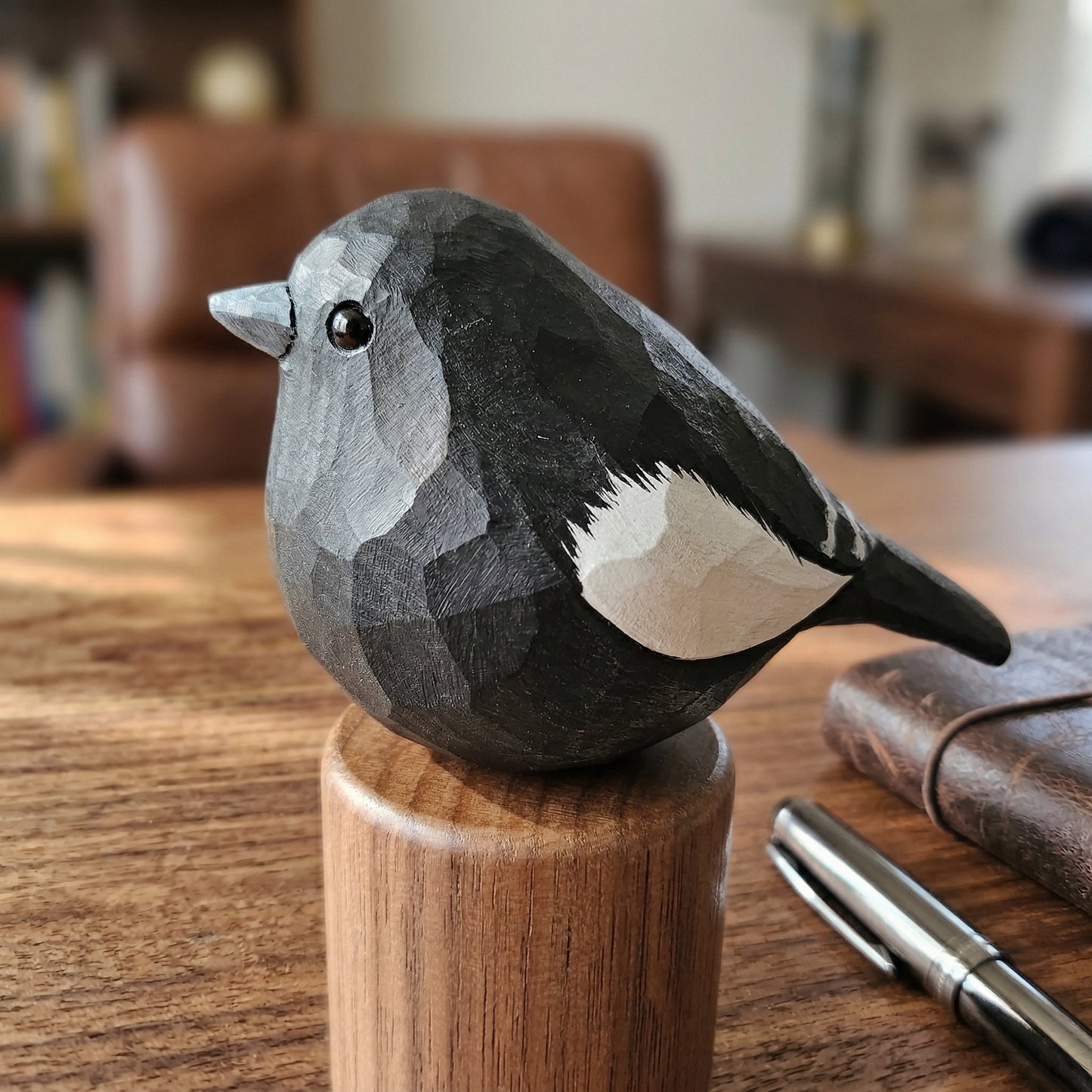 A hand-carved wooden Lark bunting figurine with a faceted geometric pattern, painted in black, gray, and white, perched on a cylindrical wooden stand on a wooden table.