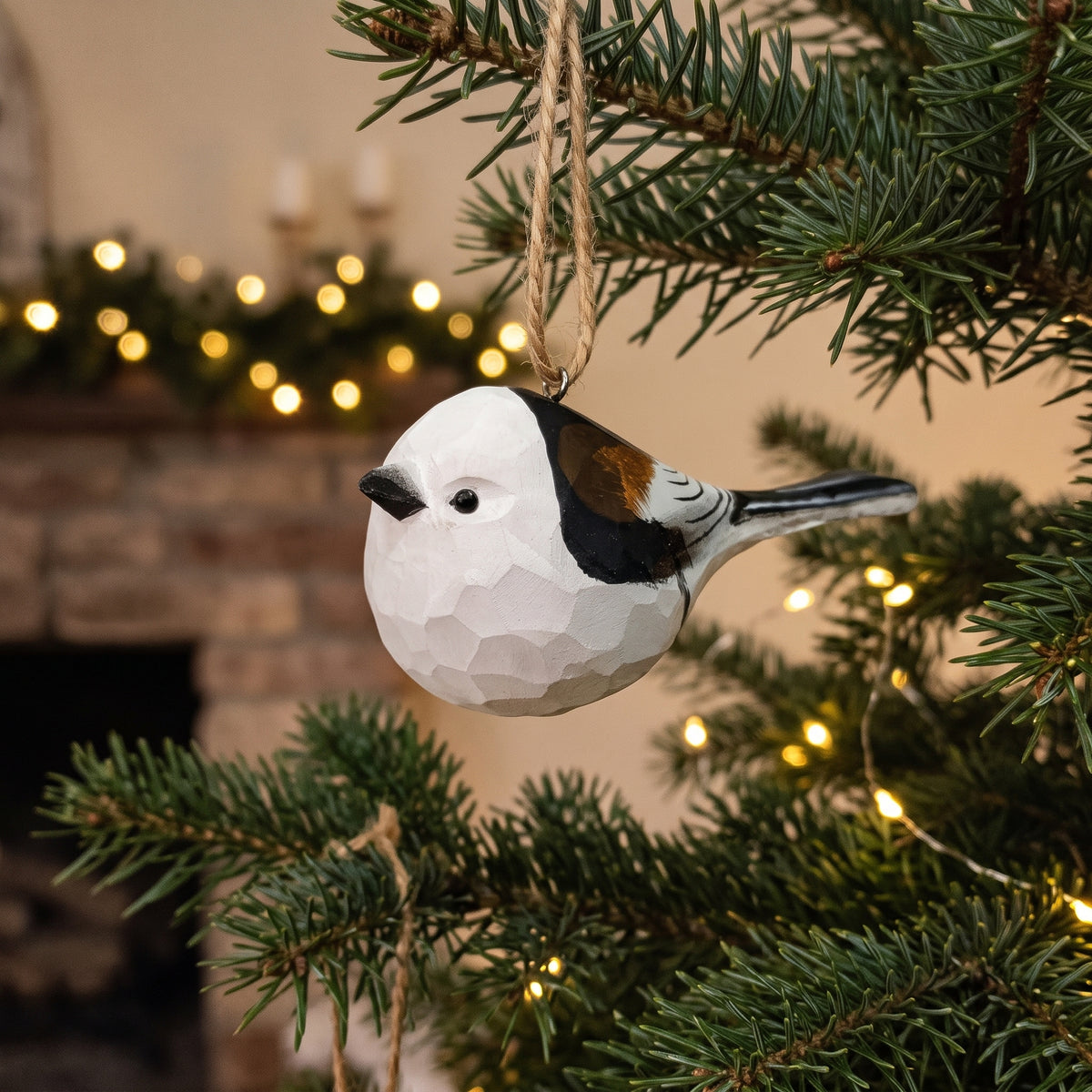 A Long-tailed tit-shaped Christmas ornament with a faceted geometric texture hangs from a tree branch. The ornament is white with black, brown, and gray details, set against a warm, blurred holiday background with lights and a fireplace.