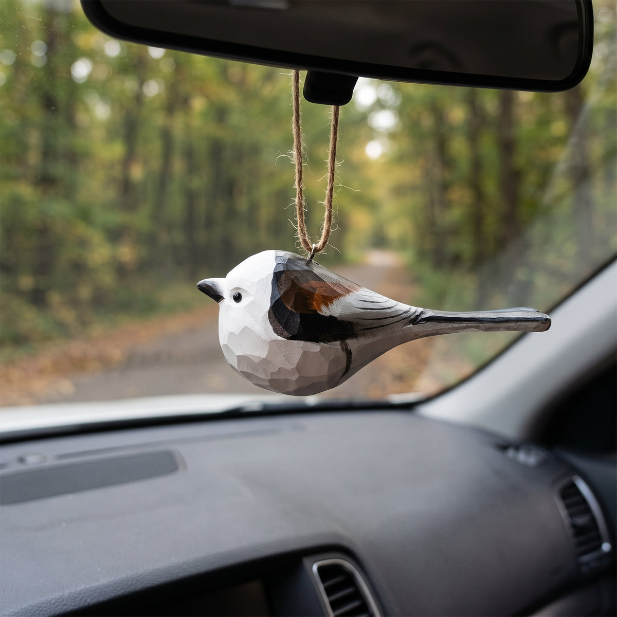 A faceted Long-tailed tit-shaped car air freshener or ornament hanging from a rearview mirror inside a car, with a blurred forest road visible through the windshield.