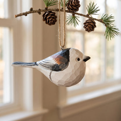 A handcrafted wooden Long-tailed tit ornament, shaped like a chickadee with a faceted geometric finish, hanging from a pine branch with pinecones in front of a softly blurred window.