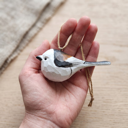 A hand holding a small, faceted Long-tailed tit ornament in white, gray, black, and brown, with a natural twine hanging loop, set against a soft beige background.