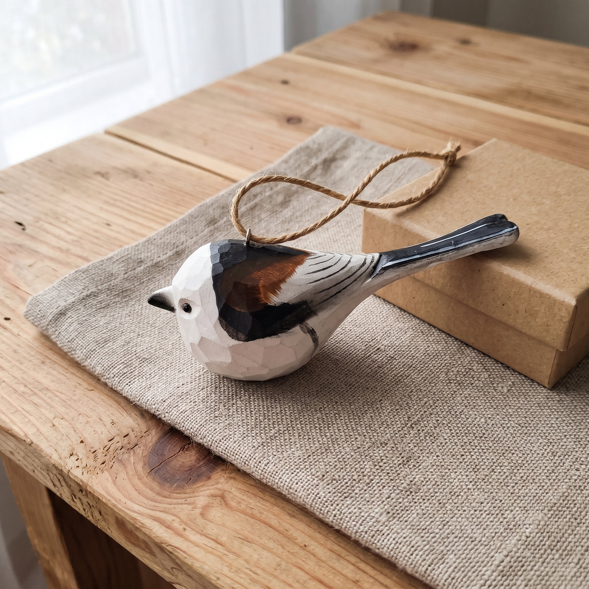 A faceted ceramic Long-tailed tit ornament, resembling a chickadee, with a hand-painted black, white, and brown pattern, hanging from a natural jute string. It is displayed on a rustic wooden table next to a brown kraft gift box and a beige linen cloth.